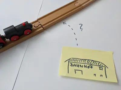 A toy train on wooden tracks and a station drawn on a post-it note. A question mark is drawn between the two.